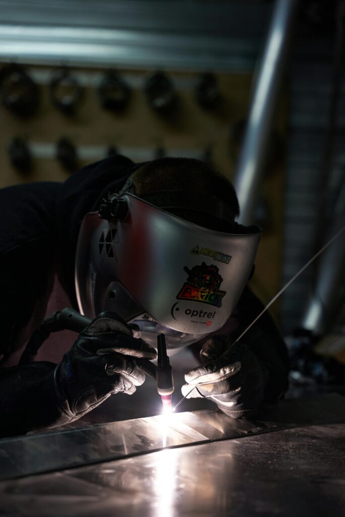 Metal worker using a TIG welding torch in a dimly lit workshop, showcasing precision and skill.